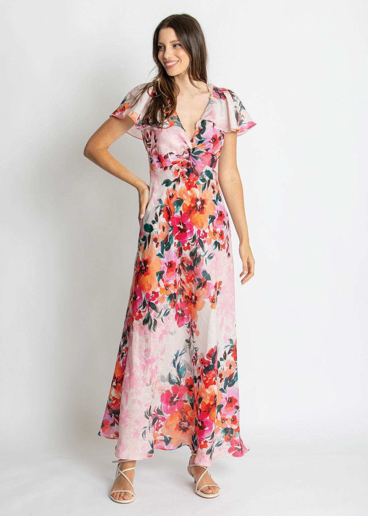A woman modeling a long floral maxi dress with flutter sleeves and a deep V-neckline, standing against a plain light gray studio background.