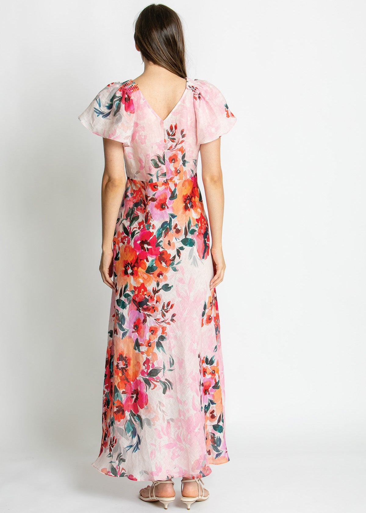 A woman viewed from the back wearing a long sheer pink dress with a large multicolor floral print and flutter sleeves, standing against a plain light gray studio background.