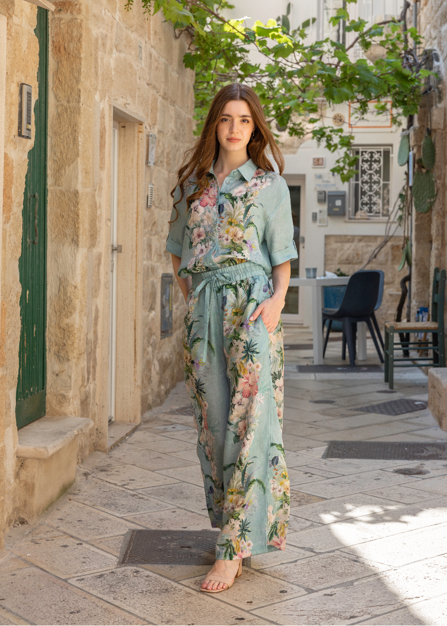 A woman stands barefoot in a sunlit stone alleyway wearing a matching light blue floral pajama set with a short-sleeve button-front top and wide-leg pants.