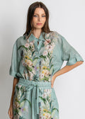 A woman modeling a matching two-piece set consisting of a short-sleeve button-up shirt and coordinating drawstring pants, both in a light sage green fabric with a colorful tropical floral and bird print.
