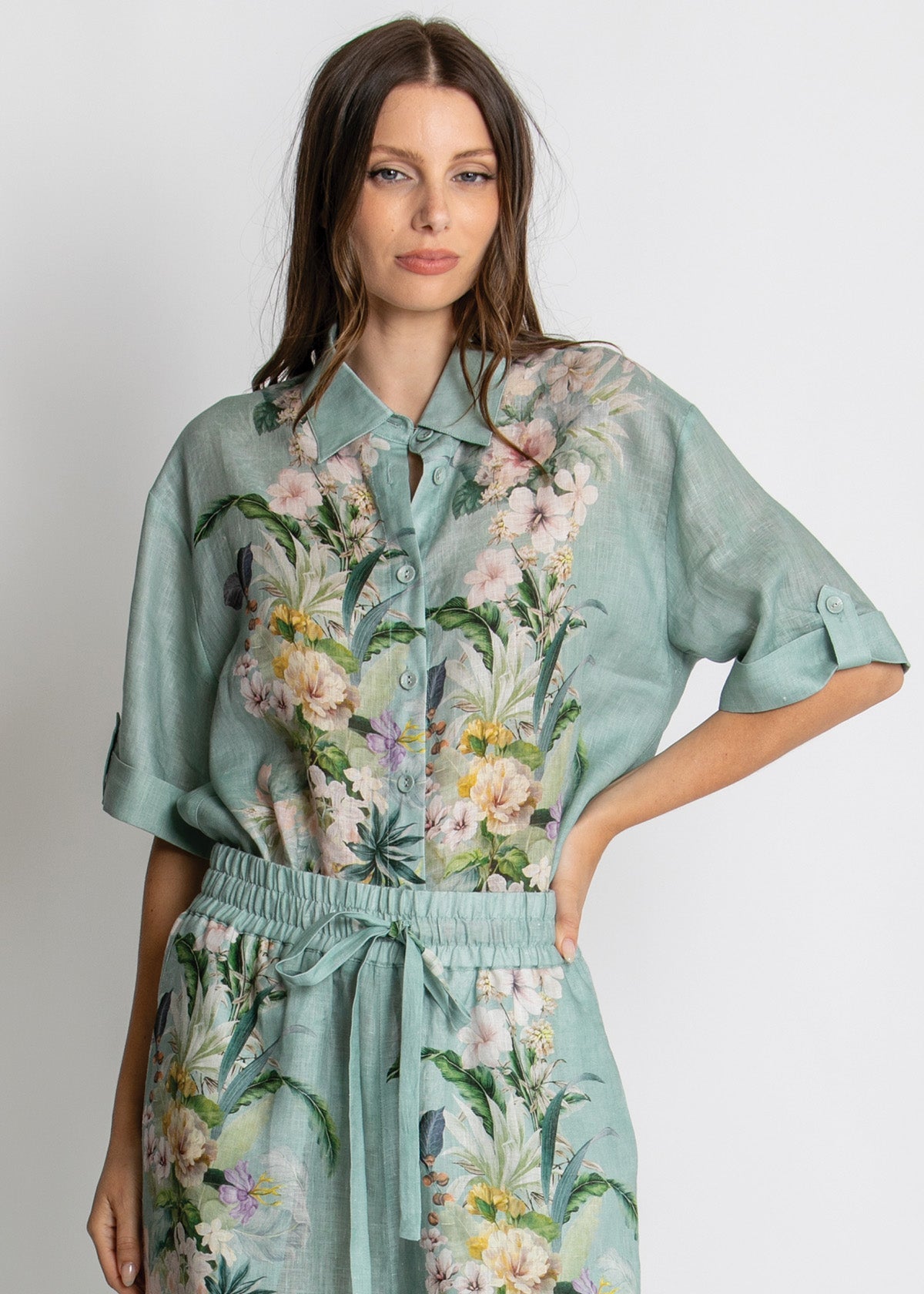 A woman modeling a matching two-piece set consisting of a short-sleeve button-up shirt and coordinating drawstring pants, both in a light sage green fabric with a colorful tropical floral and bird print.