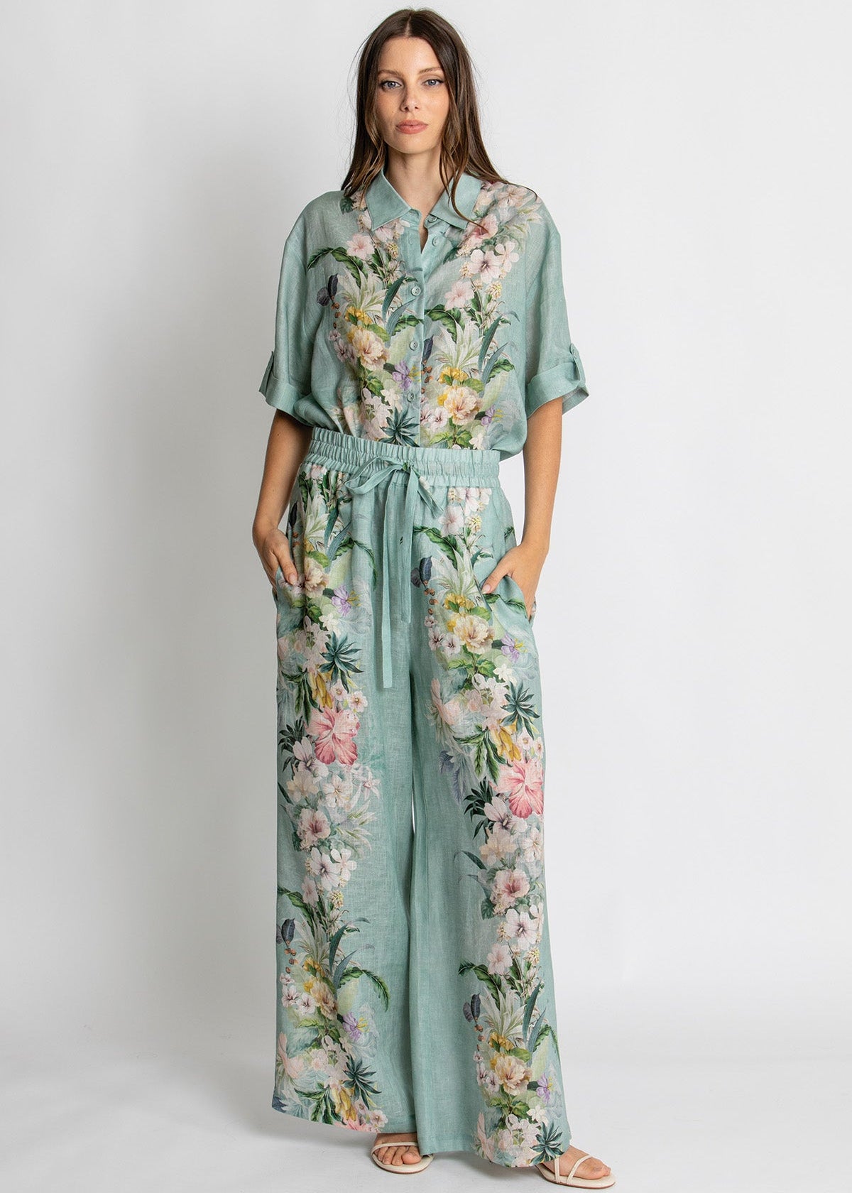 A woman modeling a mint green two-piece set featuring a short-sleeve button-up shirt and matching wide-leg pants, both decorated with a colorful tropical floral and palm leaf print. She is standing against a plain light gray studio background.