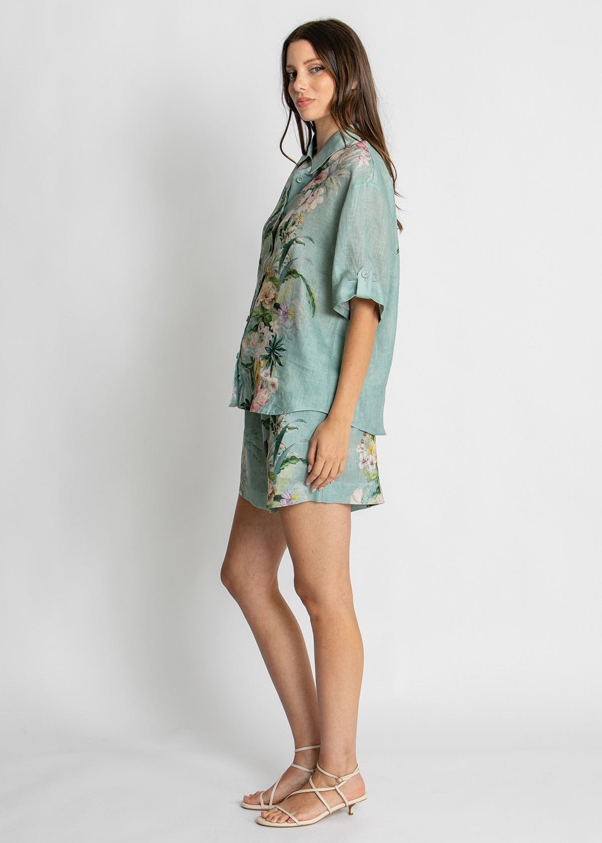 A woman modeling a matching light blue-green short-sleeve button-up shirt and shorts set with a floral print, paired with beige strappy sandals, against a plain light gray studio background.