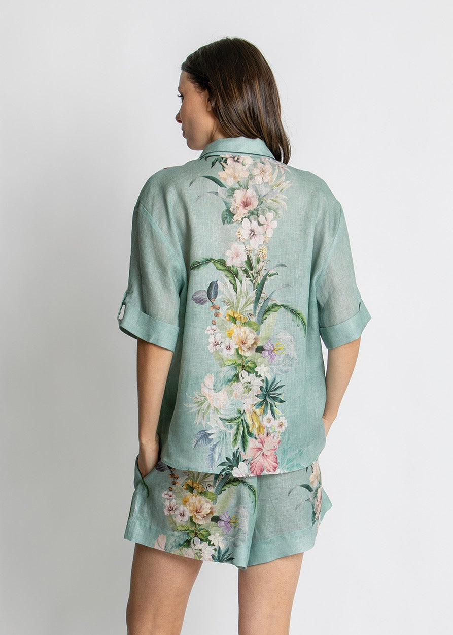 A woman shown from the back wearing a mint green short-sleeve button-up shirt and matching shorts set with a large multicolor floral print, paired with beige strappy sandals, against a light gray studio background.