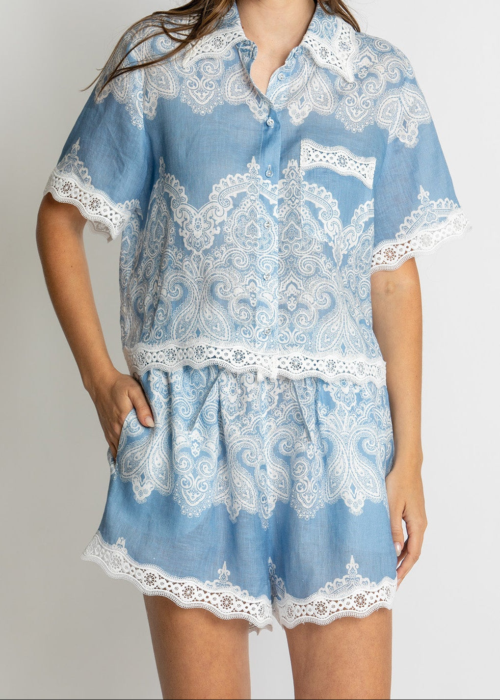 A woman modeling a light blue two-piece pajama set with white lace trim, featuring a short-sleeve button-up top and matching shorts.