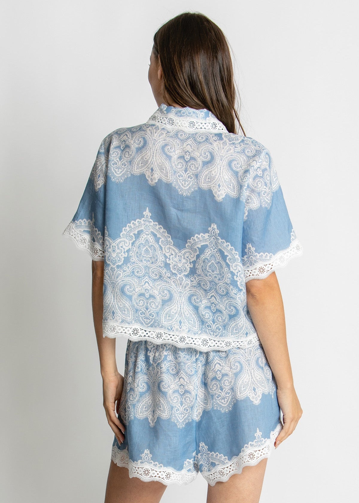 A woman shown from the back wearing a light blue two-piece set with white lace trim, consisting of a short-sleeve collared top and matching shorts, against a plain light gray background.