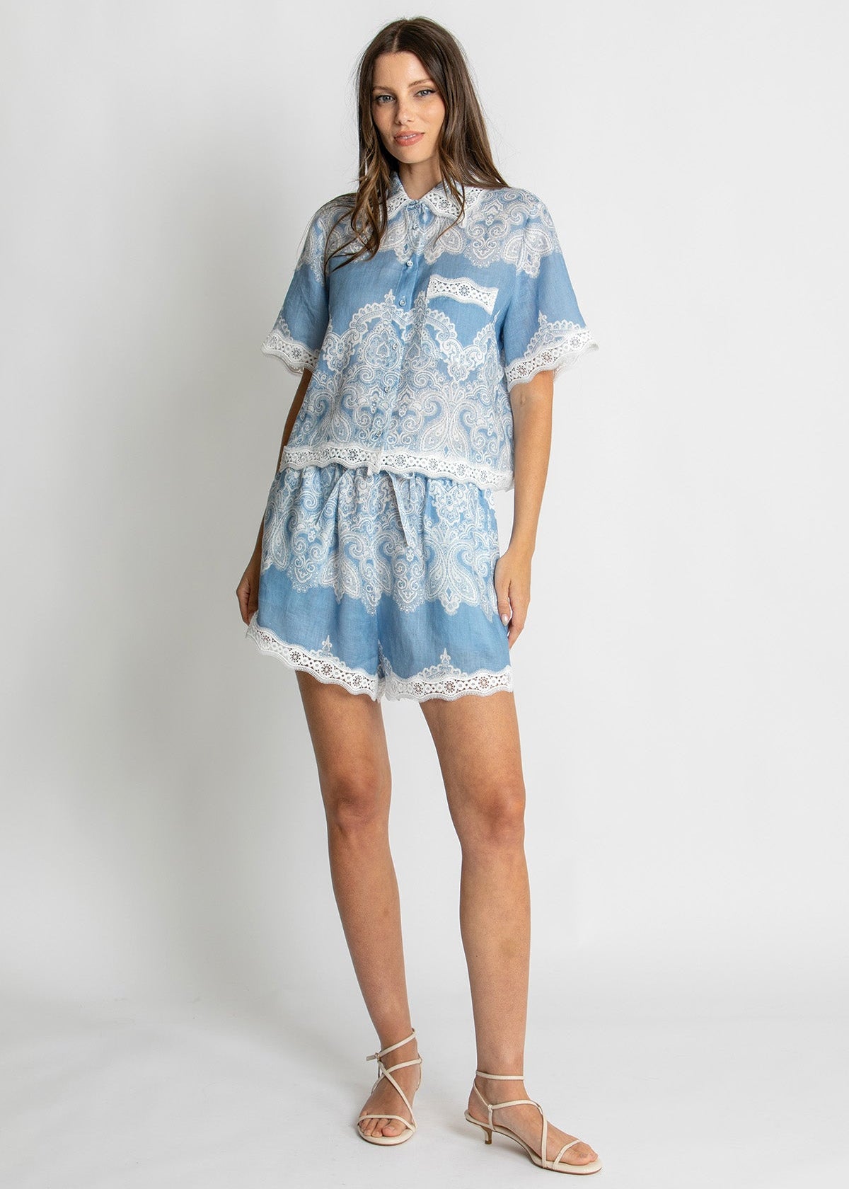 A woman modeling a light blue and white lace two-piece set consisting of a short-sleeve button-up top and matching shorts, paired with white strappy sandals, against a plain light gray studio background.