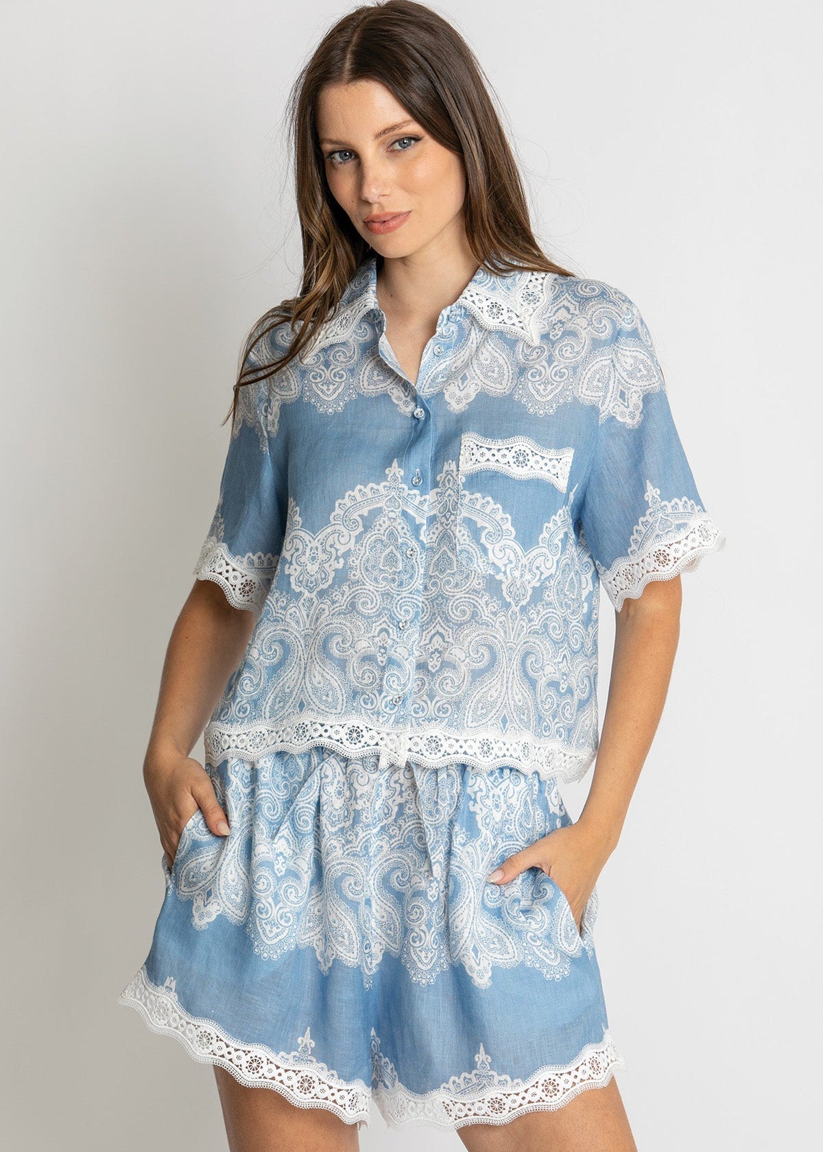 A woman modeling a light blue and white lace pajama set consisting of a short-sleeve button-up shirt and matching shorts, photographed against a neutral light gray background.