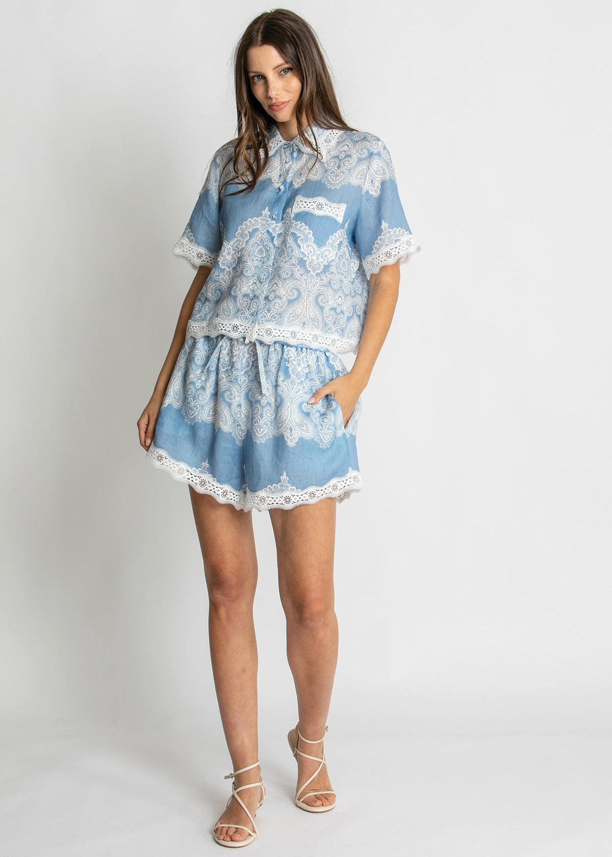 A woman modeling a light blue and white lace-patterned two-piece set consisting of a short-sleeve button-up top and a matching mini skirt, paired with beige strappy sandals against a plain light gray studio background.