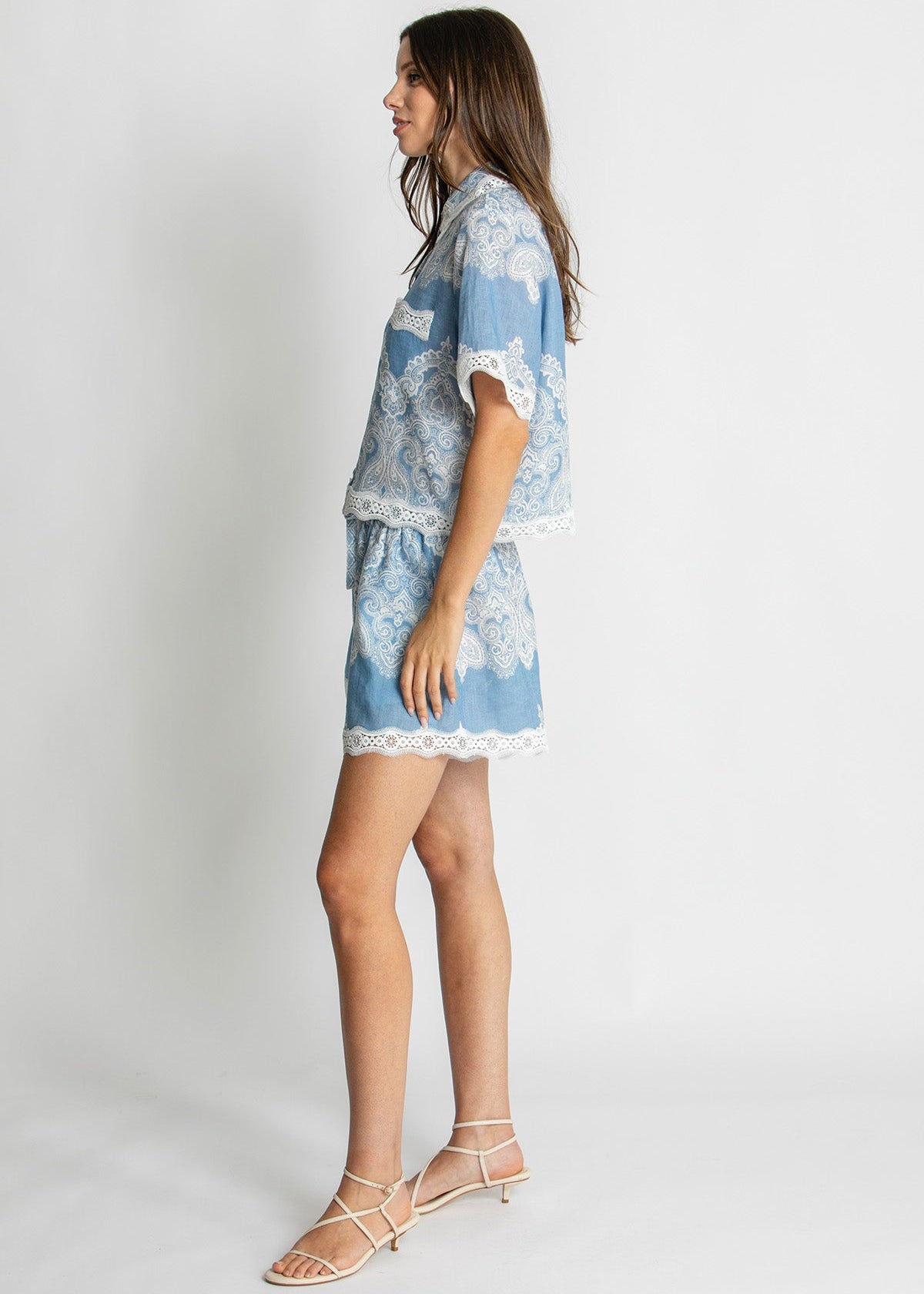 A woman modeling a light blue and white patterned two-piece set consisting of a short-sleeve top and matching shorts, styled with white strappy sandals against a plain light gray studio background.