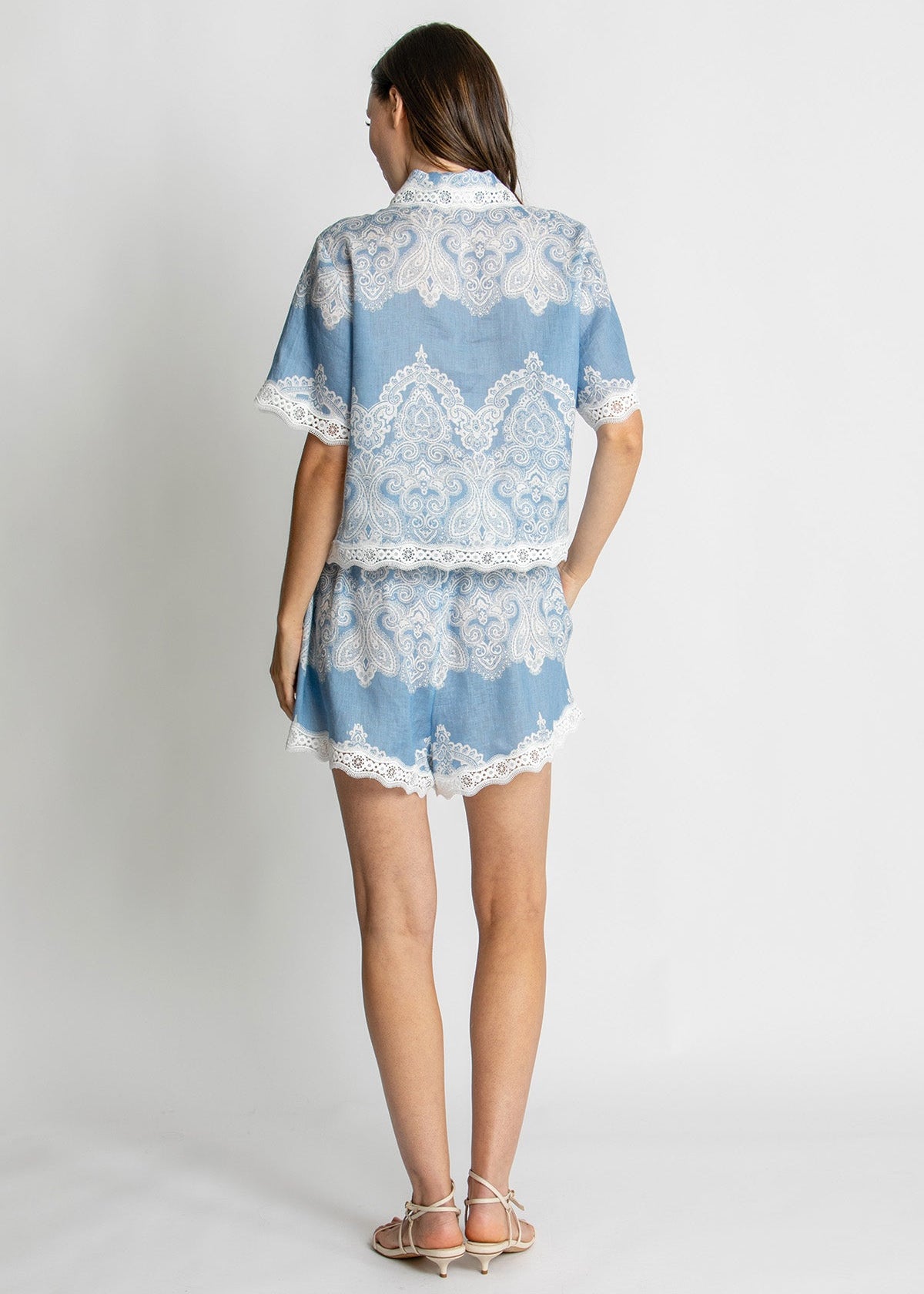 A woman shown from the back wearing a light blue two-piece set with white lace trim, consisting of a short-sleeve collared top and matching shorts, against a plain light gray studio background.