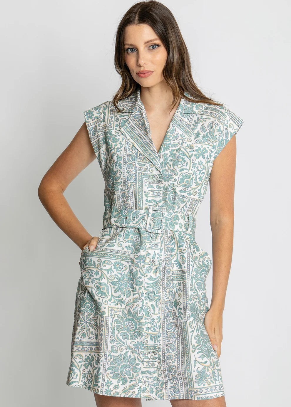 A woman modeling a sleeveless wrap-style dress with a matching waist tie, featuring a light blue and white paisley pattern, set against a plain light gray studio background.