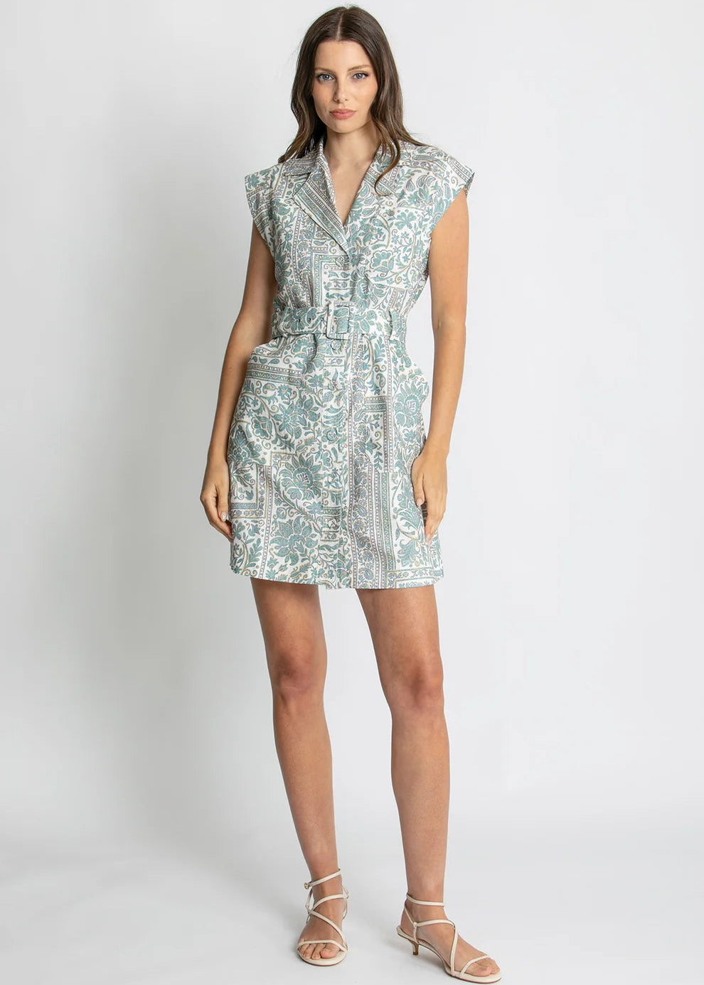 A woman modeling a sleeveless mini dress with a white base and a light blue botanical pattern, styled with beige strappy high-heeled sandals against a light gray studio background.