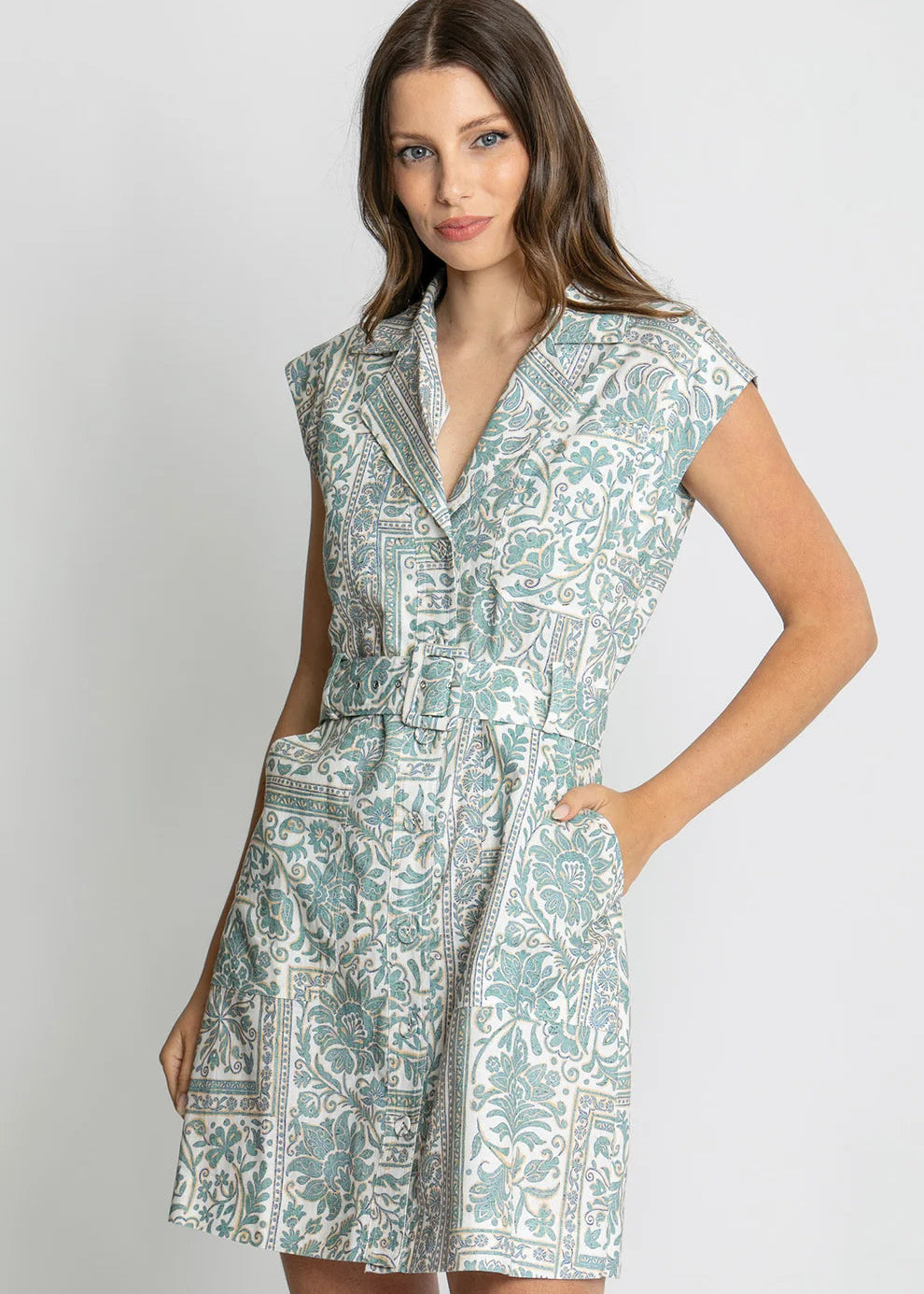 A woman modeling a short, sleeveless wrap-style dress with a V-neck, belt, and side pockets. The dress features an intricate paisley pattern in sage green and white.