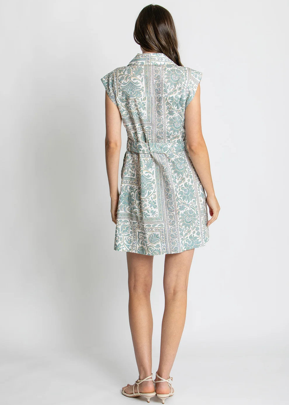 A woman viewed from the back wearing a sleeveless collared dress with a light blue and white ornate pattern and a matching tie belt, paired with beige strappy heels, against a light gray studio background.