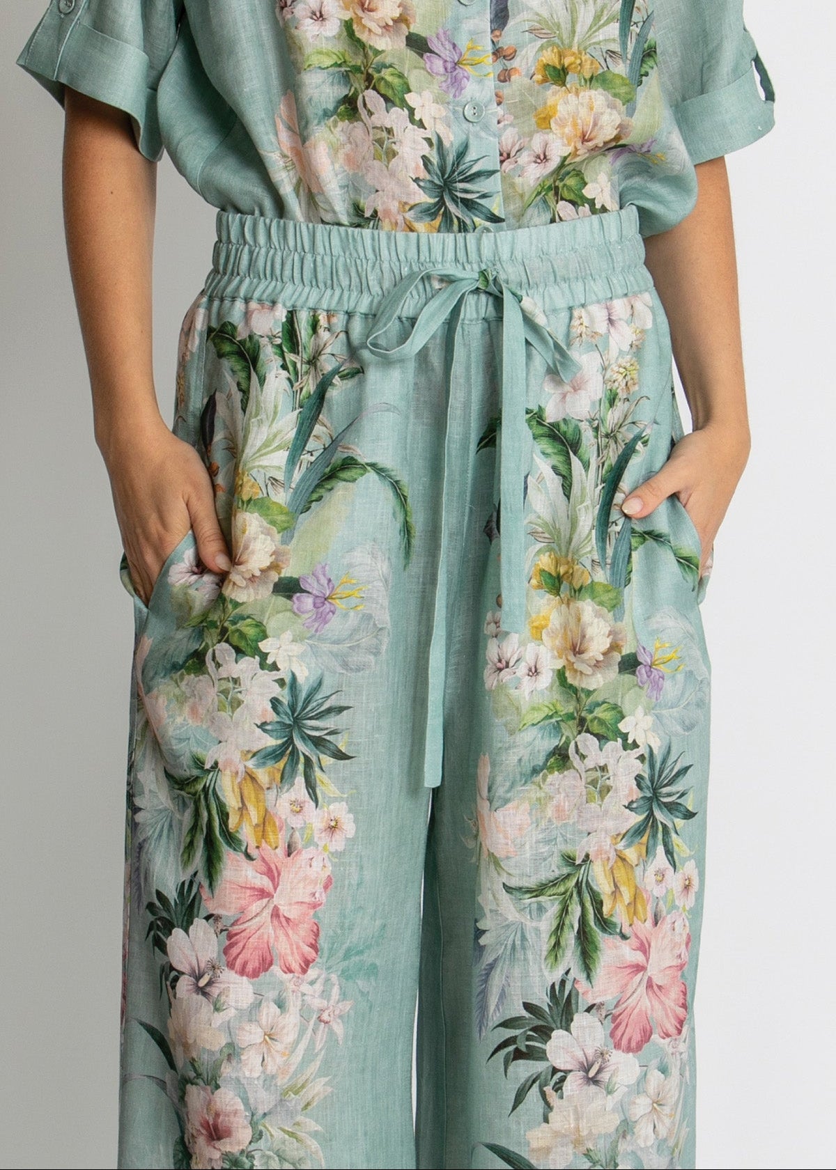 A person wearing a mint green floral jumpsuit with a tie waist and side pockets, shown from the chest down against a plain light background.
