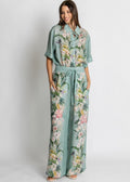 A woman modeling a matching two-piece set consisting of a short-sleeve collared top and wide-leg pants, both in a sage green color with a vibrant multicolor floral print, set against a plain light gray background.