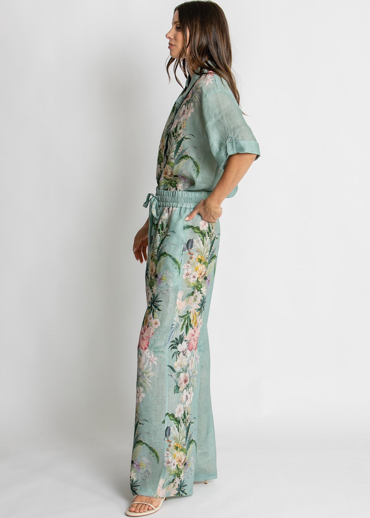 A woman modeling a sage green floral print jumpsuit with short sleeves, a tie waist, and wide-leg pants, shown from a side profile against a light gray studio background.