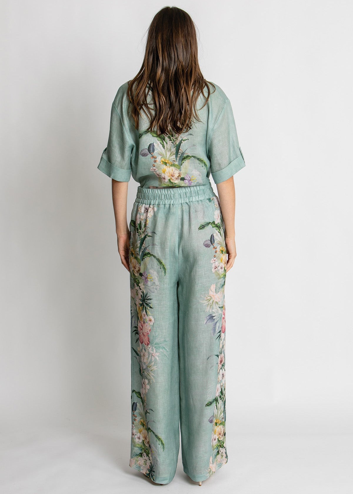 A woman viewed from behind wearing a sage green jumpsuit with a floral print, standing against a plain light gray studio background.