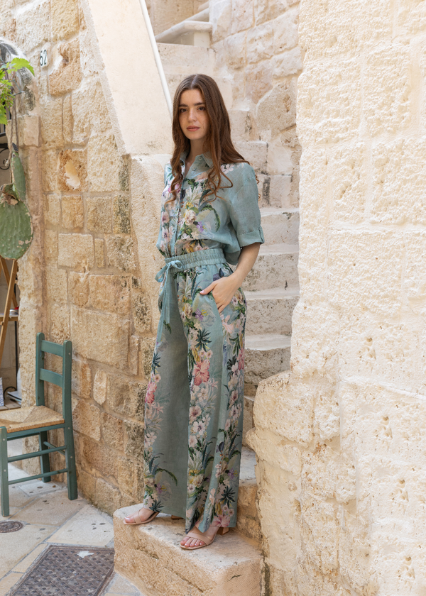 A woman stands on outdoor stone steps wearing a matching two-piece jumpsuit in sage green with a colorful tropical floral print. The set includes a short-sleeve collared top and wide-leg trousers with a tie waist.