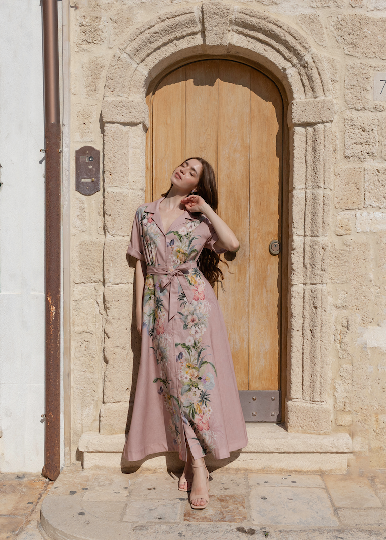 A woman wearing a dusty pink floral maxi dress stands in front of a rustic stone building with a wooden door.