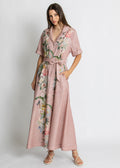 A woman modeling a dusty pink short-sleeve maxi dress with a floral print and a matching tie belt, set against a plain light gray studio background.