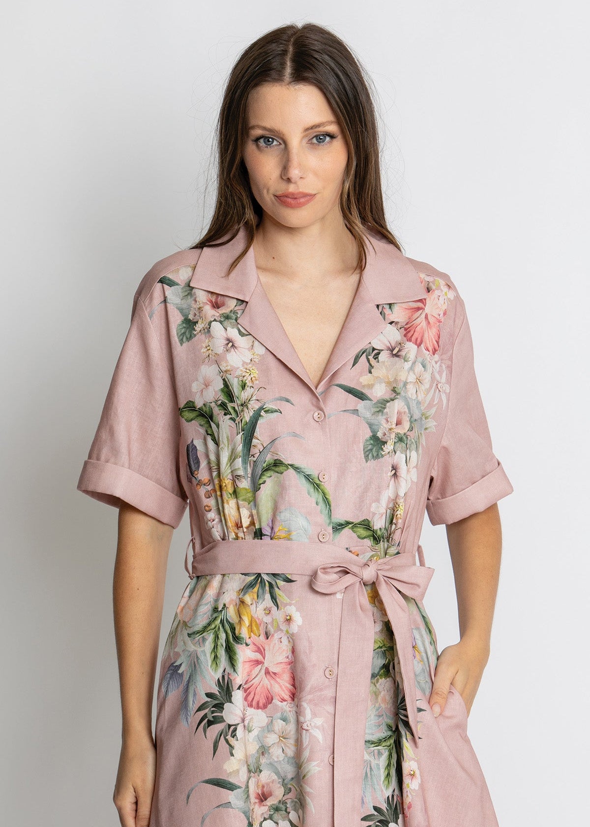A woman wearing a dusty pink short-sleeve shirt dress with a floral print, featuring a matching tie belt, photographed against a light gray studio background.