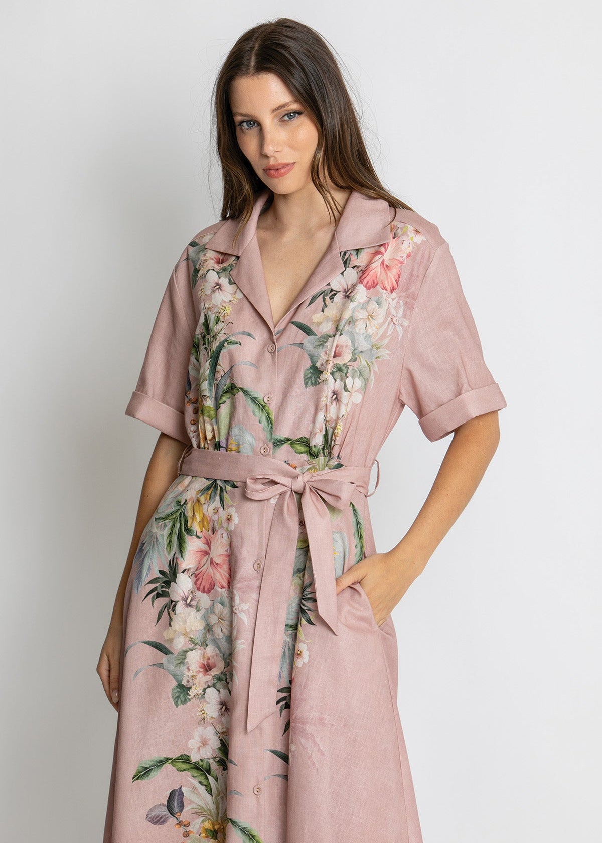 A woman modeling a dusty pink short-sleeve wrap dress with a floral print and matching tie belt, set against a plain light gray studio background.