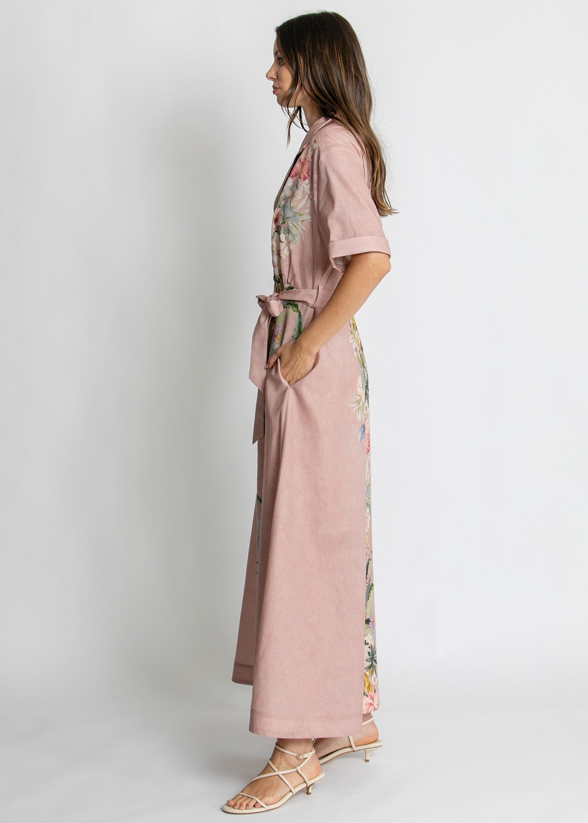 A woman modeling a dusty pink wrap maxi dress with short sleeves, a matching tie belt, and multicolor floral embroidery along the side and front, paired with beige strappy sandals against a light gray studio background.