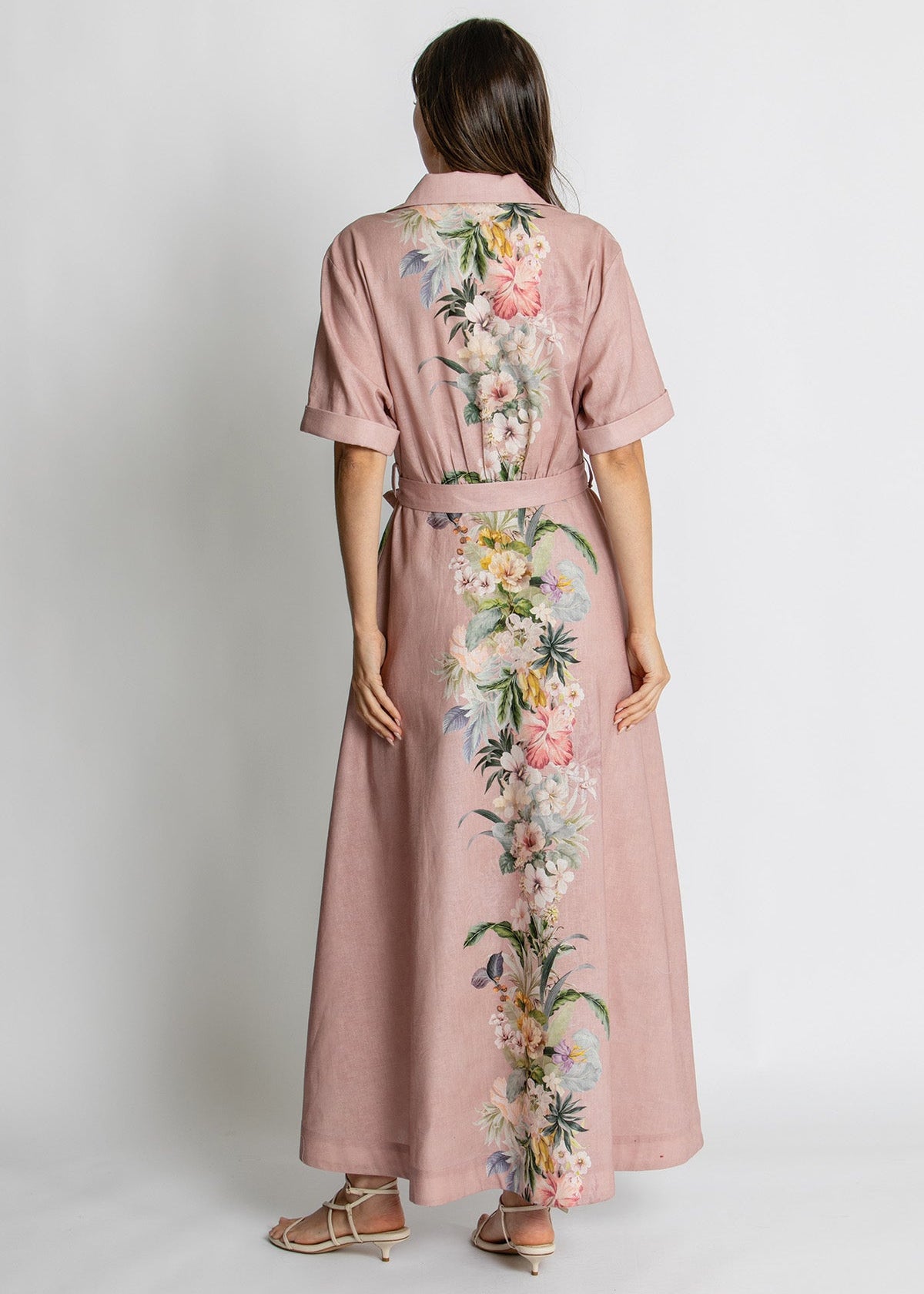 A woman viewed from the back wearing a dusty pink short-sleeve maxi dress with a vertical multicolor floral print down the center back and sides, paired with tan heeled sandals against a light gray studio background.