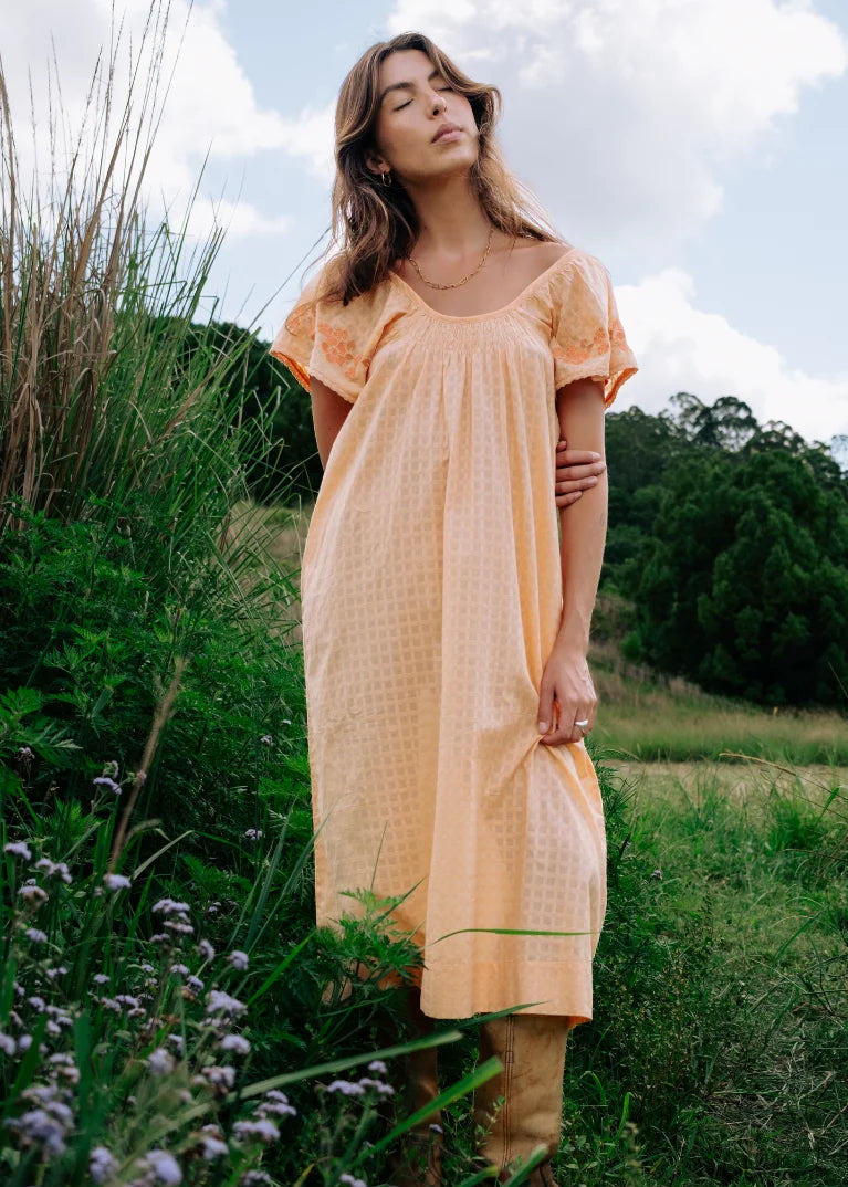 A woman wearing a light peach short-sleeve midi dress with a subtle textured pattern, standing outdoors in a grassy field under a cloudy sky.