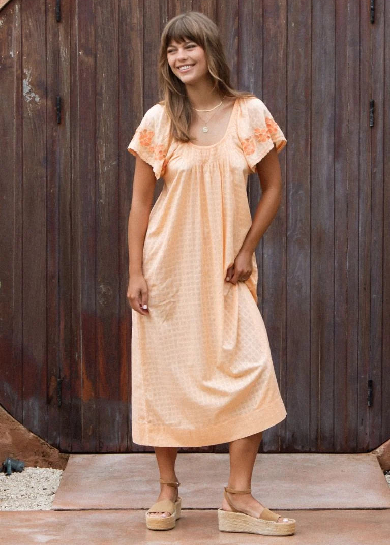 A woman wearing a peach-colored midi dress with short flutter sleeves and orange floral embroidery on the cuffs, standing in front of a rustic dark brown wooden door.