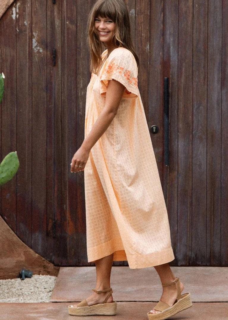 A woman wearing a light peach midi dress with short flutter sleeves and orange floral embroidery on the cuffs, standing in front of a rustic dark brown wooden door.