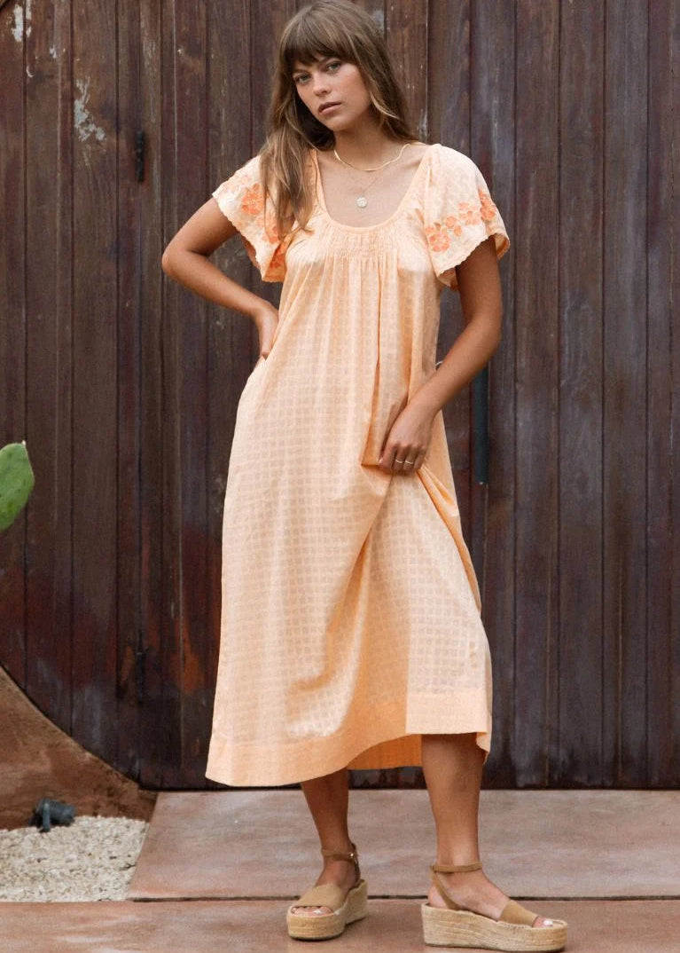 A woman wearing a light peach midi dress with short flutter sleeves and floral embroidery on the shoulders, standing in front of a dark wooden door.