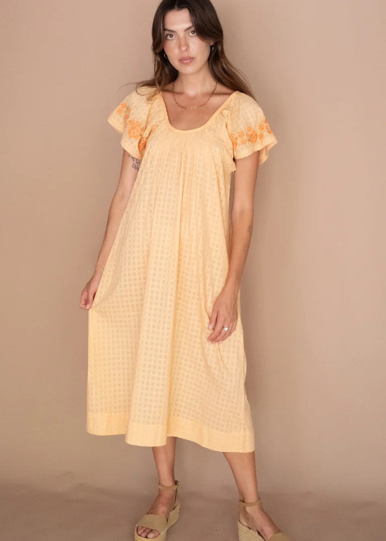 A woman modeling a pale yellow midi dress with a subtle grid pattern, short flutter sleeves with orange embroidery, and tan wedge sandals, set against a solid beige studio backdrop.