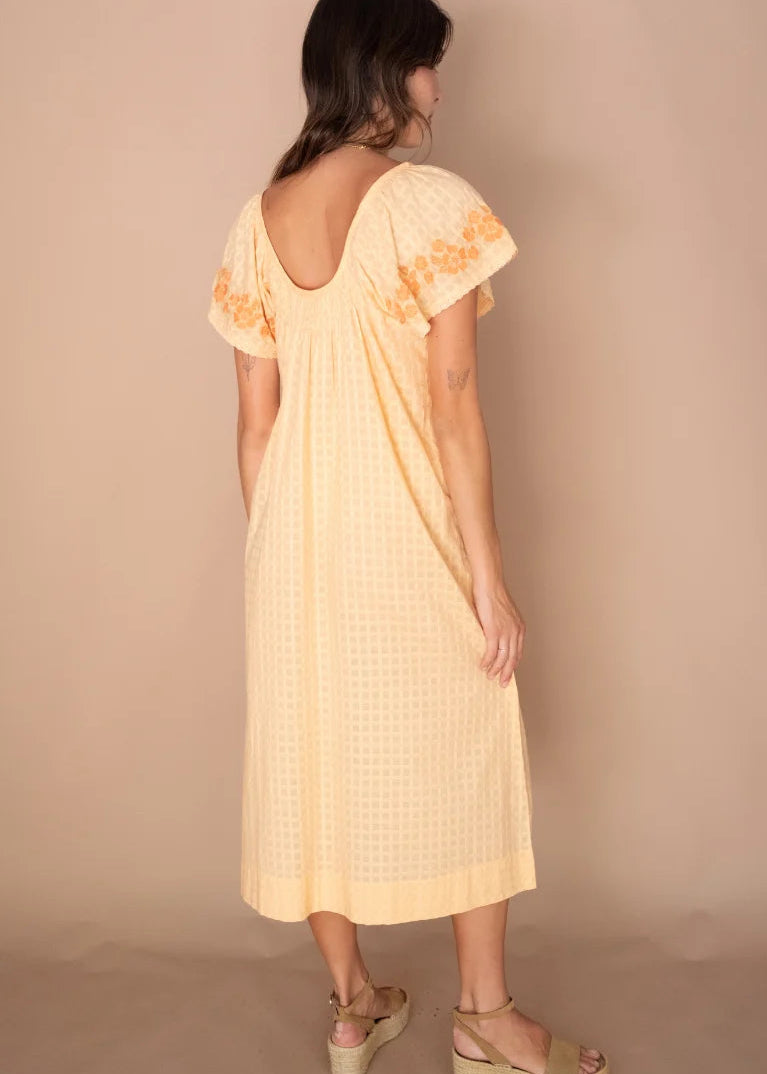 A woman shown from the back wearing a pale yellow midi dress with short flutter sleeves, embroidered details on the shoulders, and a small gingham pattern. She is wearing beige wedge sandals against a solid beige studio backdrop.