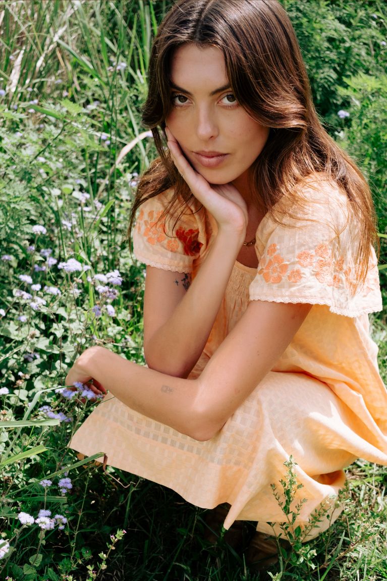 A woman with long brown hair crouches in a grassy field of green plants and small purple flowers, wearing a light peach-orange dress with lace trim.
