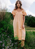 A woman stands outdoors in a grassy field wearing a loose, short-sleeve peach-colored dress with a subtle textured pattern and tan boots.