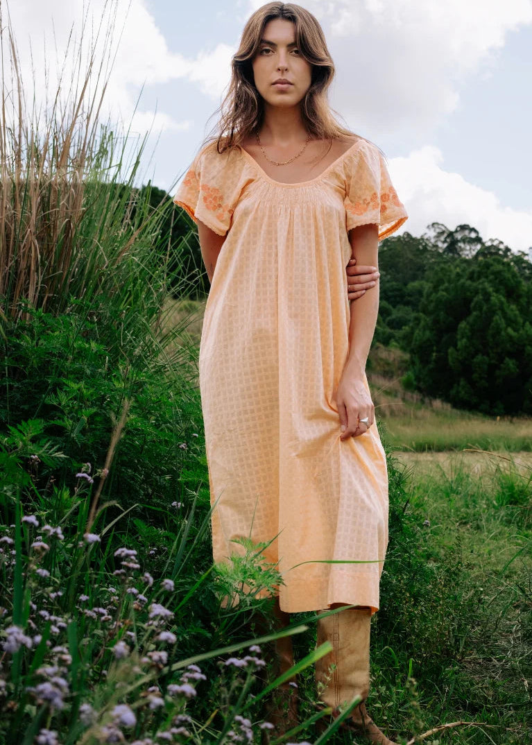 A woman stands outdoors in a grassy field wearing a loose, short-sleeve peach-colored dress with a subtle textured pattern and tan boots.