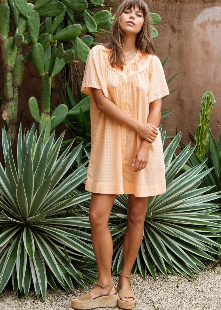 A woman wearing a short-sleeve peach-colored cotton dress and beige platform sandals, standing outdoors in front of a terracotta wall and green cacti and agaves.
