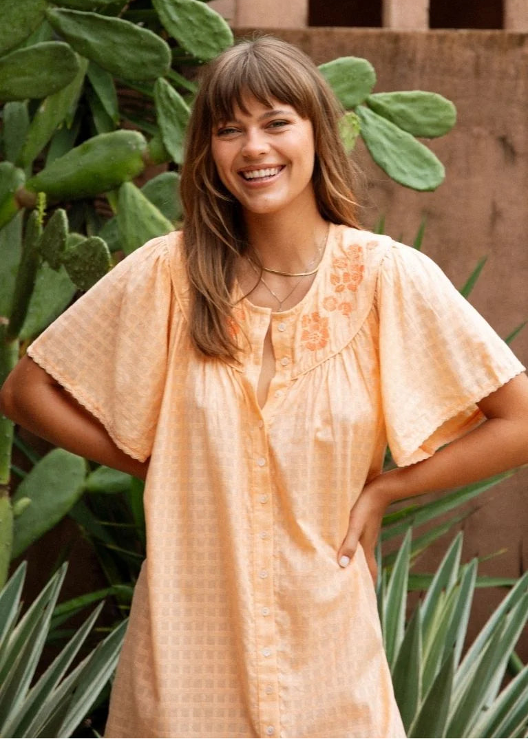 A woman smiling outdoors wearing a loose, short-sleeve peach-colored tunic top with embroidered details on the chest. The background features green cacti and a brown wooden wall.