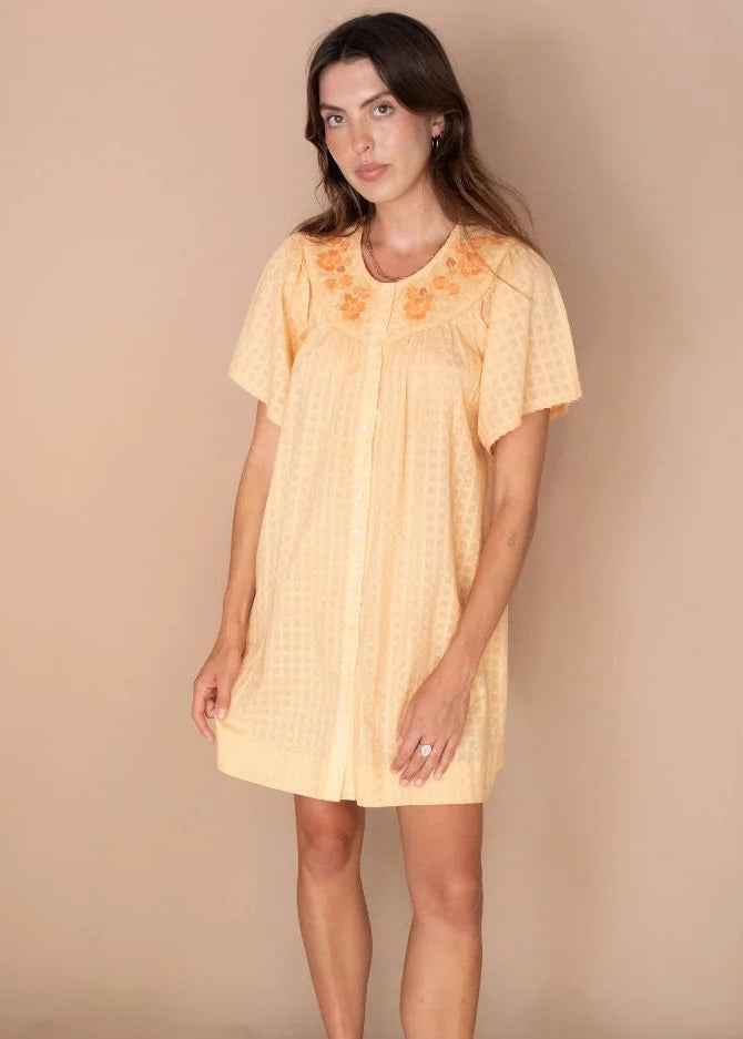 A woman modeling a pale yellow short-sleeve button-front dress with orange floral embroidery around the neckline, paired with beige wedge sandals, against a solid beige studio background.