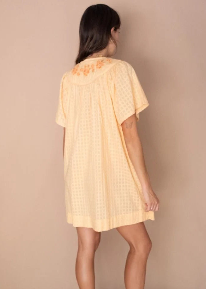 A woman shown from the back wearing a pale yellow short-sleeve tunic dress with a subtle textured pattern and orange floral embroidery on the upper back, paired with beige platform sandals against a solid beige studio backdrop.