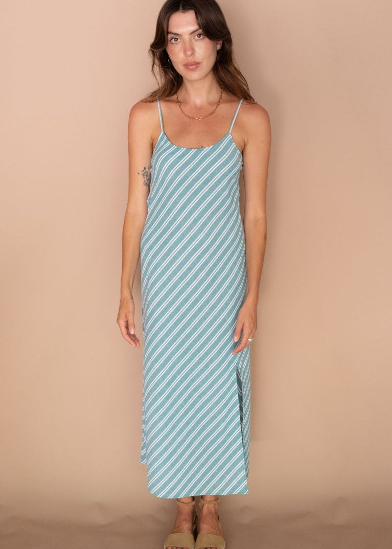 A woman modeling a light blue and white diagonal striped spaghetti-strap maxi dress against a solid beige studio backdrop.