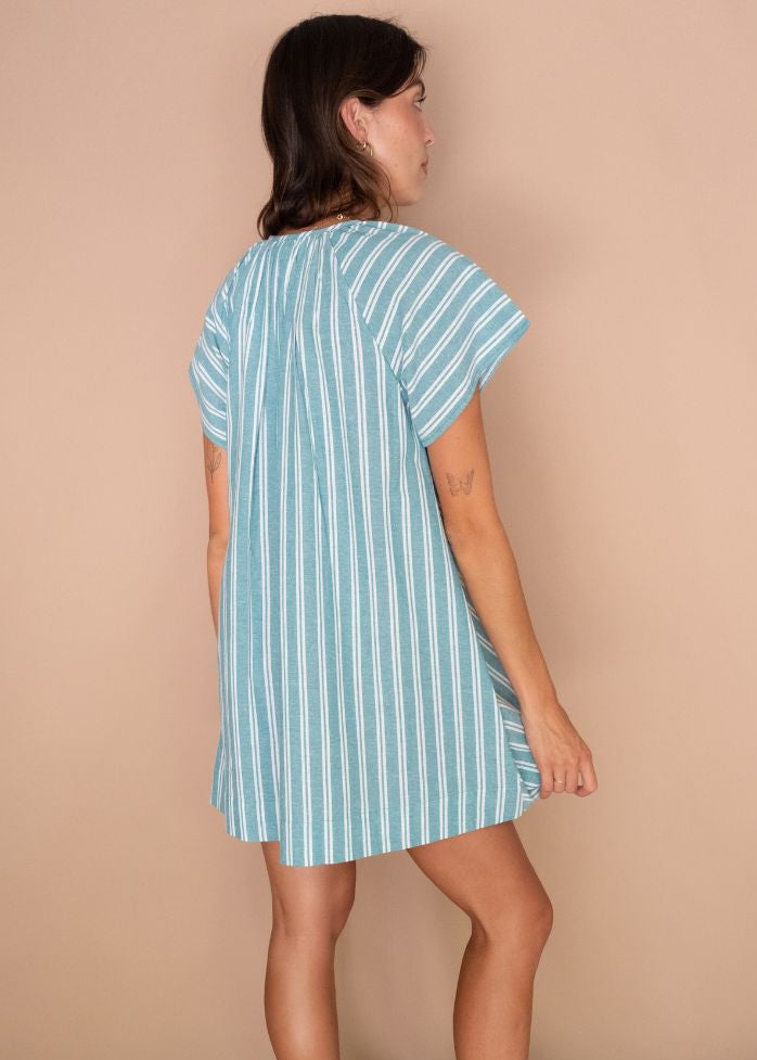 A woman wearing a light blue and white vertically striped short-sleeve tunic dress, shown from a side-back angle against a solid beige studio backdrop.