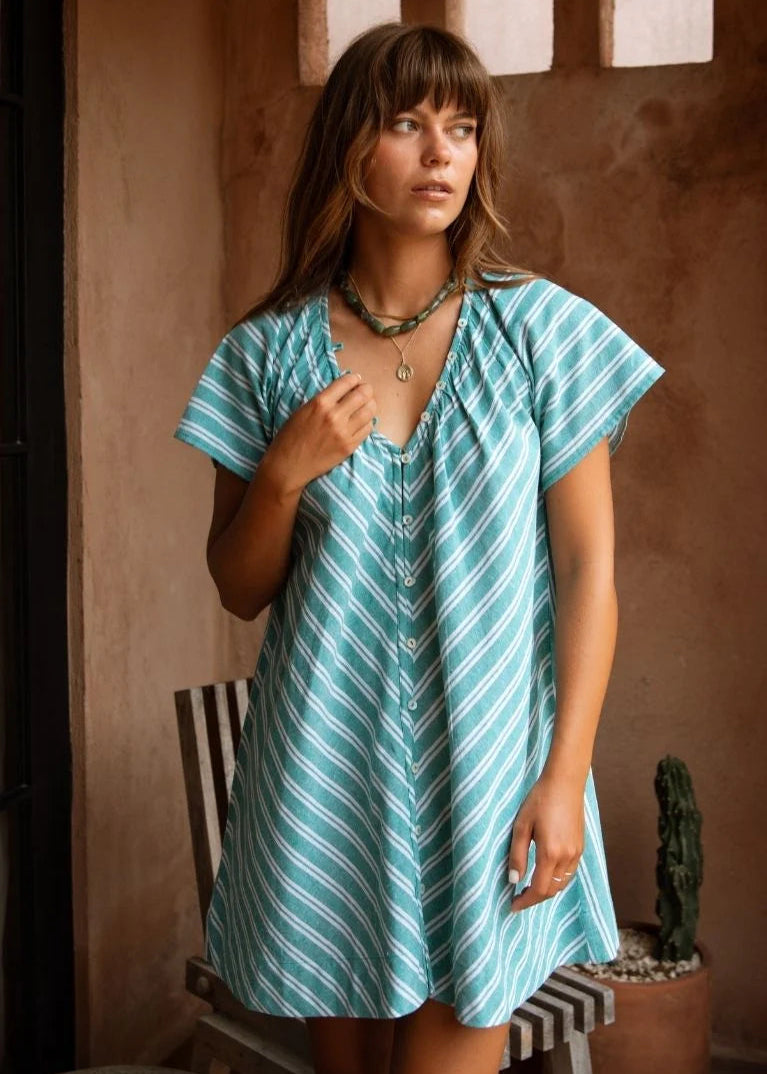 A woman wearing a short-sleeve, V-neck dress with diagonal teal and white stripes, standing in a rustic indoor setting with a textured brown wall and a striped chair.