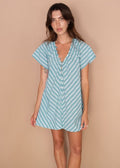 A woman modeling a light blue and white vertically striped short-sleeve mini dress with a ruffled V-neckline and button front, standing against a solid beige studio backdrop.