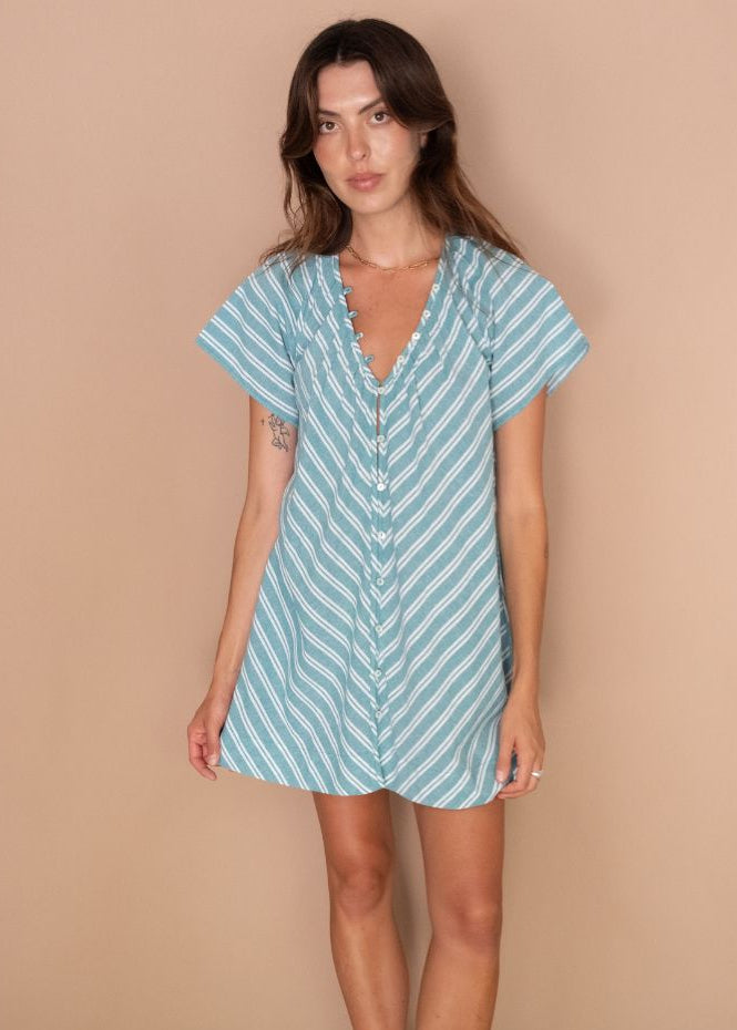 A woman modeling a light blue and white vertically striped short-sleeve mini dress with a ruffled V-neckline and button front, standing against a solid beige studio backdrop.