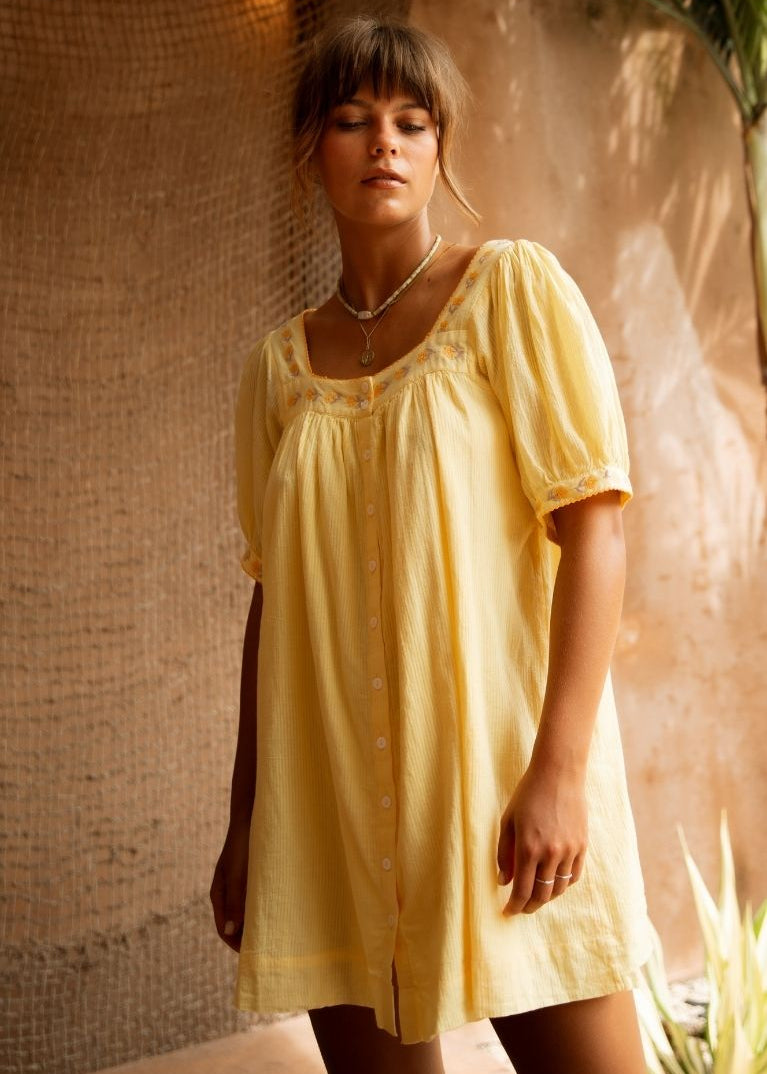 A woman wearing a pale yellow short-sleeve button-front dress or tunic with a square neckline and embroidered trim, standing in a warm-toned indoor setting with textured wall panels and plants.