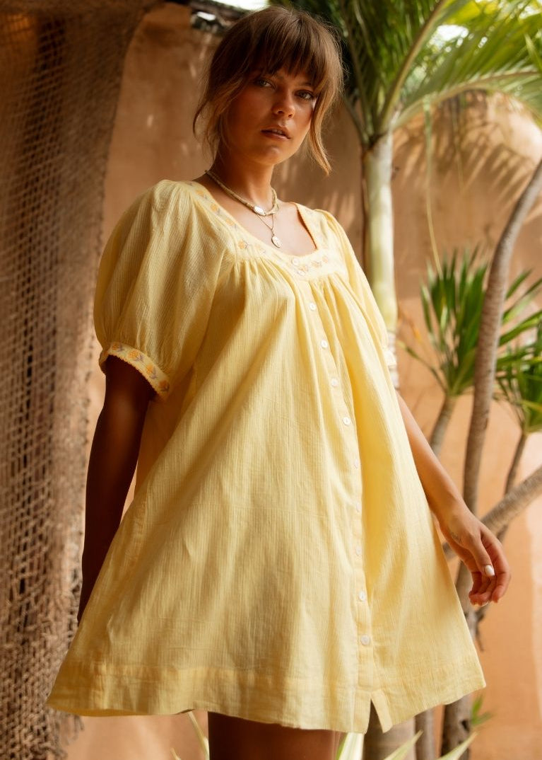 A woman wearing a pale yellow linen puff-sleeve tunic dress, standing in a rustic tropical setting with palm leaves and a warm stucco wall.